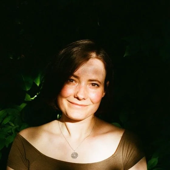 Woman with short brown hair smiling softly, illuminated by sunlight with leafy shadows in the background.