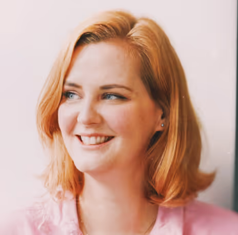 Smiling woman with shoulder-length light red hair wearing a pink shirt looking to the left.