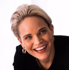 Smiling woman with short blonde hair wearing gold hoop earrings and a black top against a white background.
