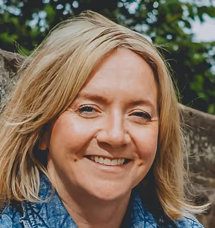 Smiling woman with blonde hair wearing a blue shirt, outdoors with greenery and stone wall background.